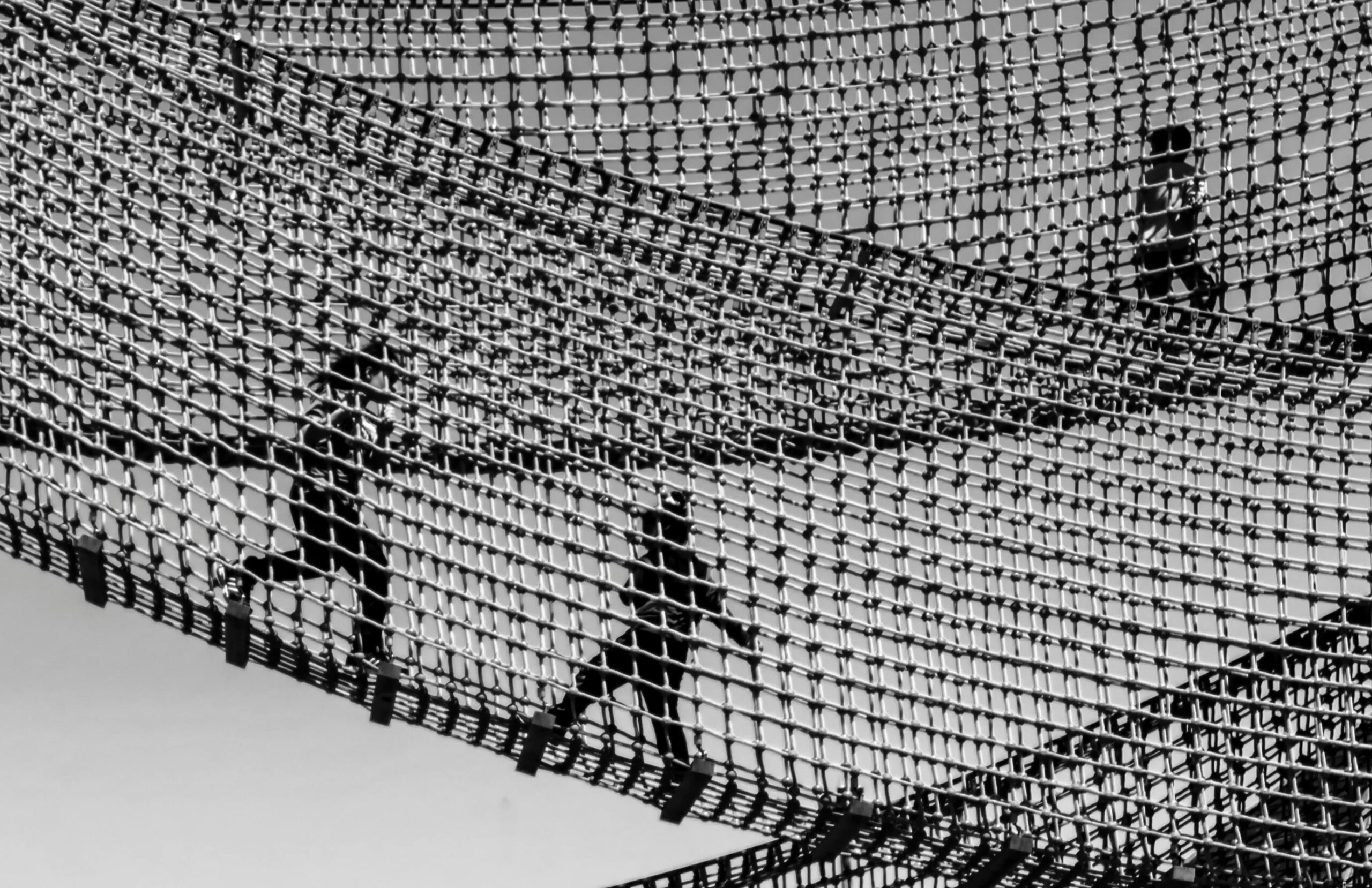 Home Silhouettes of children playing on a suspended rope bridge, captured in black and white.